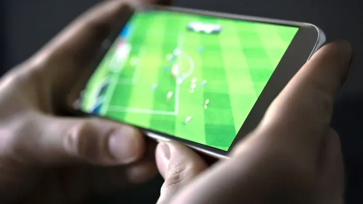 Mãos segurando o smartphone na horizontal para assistir futebol ao vivo pelo celular, com a partida na tela